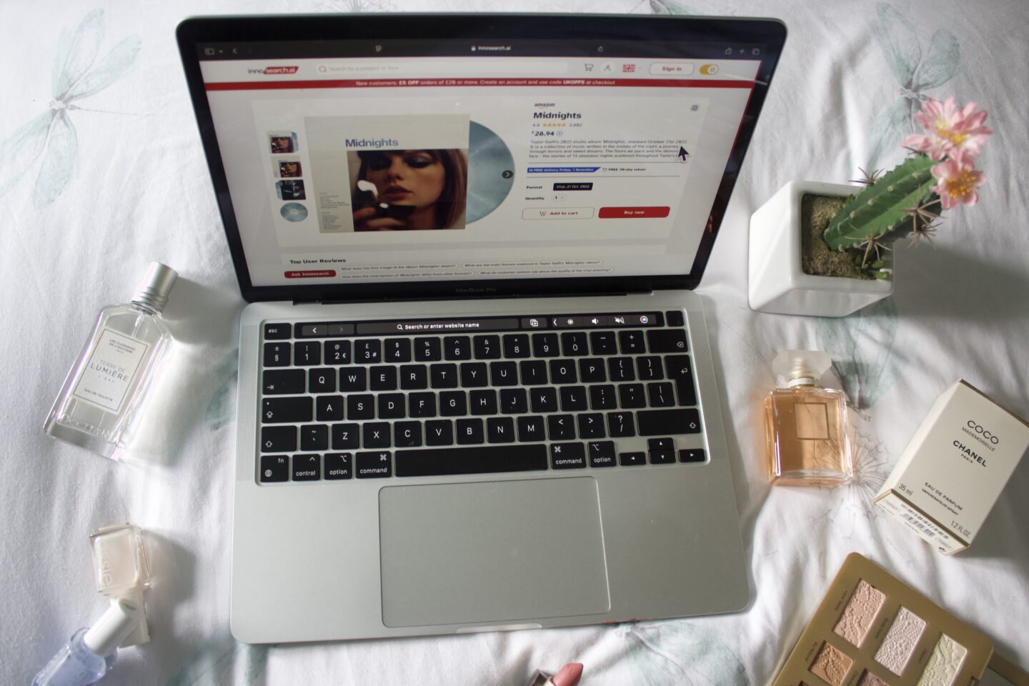 A product page for Taylor Swift's Midnights vinyl on Innosearch. The page is clean, only showing a few photos and information such as the price, a paragraph for the product description and an 'Add to cart' button. Various objects surround the laptop including a couple of perfumes, nail polishes, a fake cactus and an open eyeshadow palette.