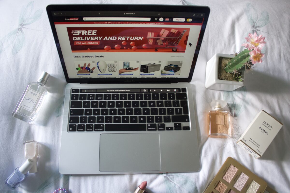 Innosearch's homepage displayed on a Macbook screen. The page shows a large red banner with white text that says 'Free delivery and returns'. There are miscellaneous items around the laptop including a couple of perfumes, a fake cactus and an open eyeshadow palette.