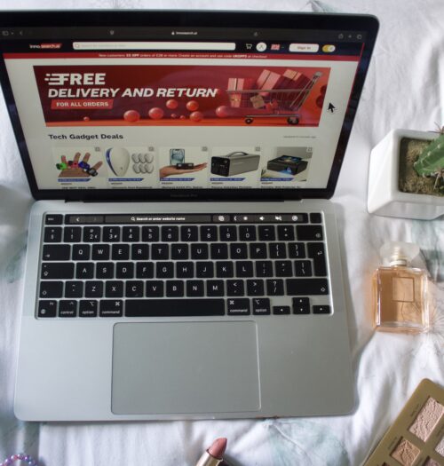 Innosearch's homepage displayed on a Macbook screen. The page shows a large red banner with white text that says 'Free delivery and returns'. There are miscellaneous items around the laptop including a couple of perfumes, a fake cactus and an open eyeshadow palette.