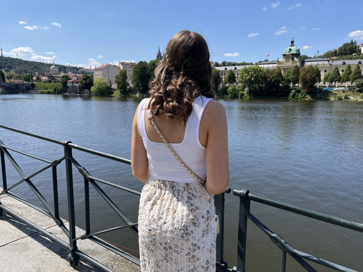 Elin looks out over the Vltava river in Prague on a sunny day. Her hair is styled into a curly half-up, half-down do with two small braids wrapping around her head to meet in the middle. She wears a white crop top which has wide straps, a beige broderie midi skirt and a white crossbody bag.