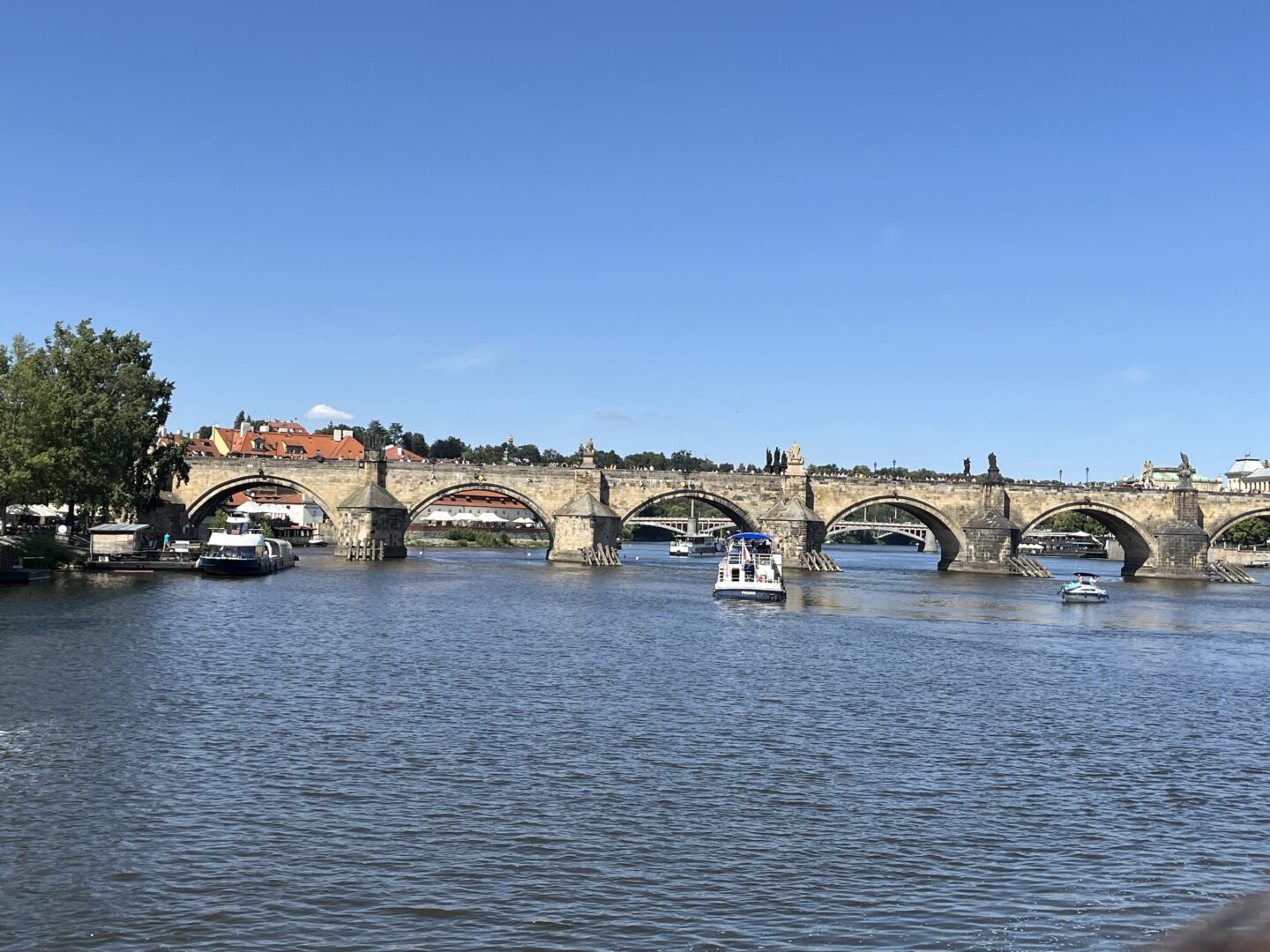 The historic Charles Bridge crossing over the Vltava river. It has several large stone arches and statues on top. Small boats move along the river towards and away from the bridge and the sky is clear blue over head.
