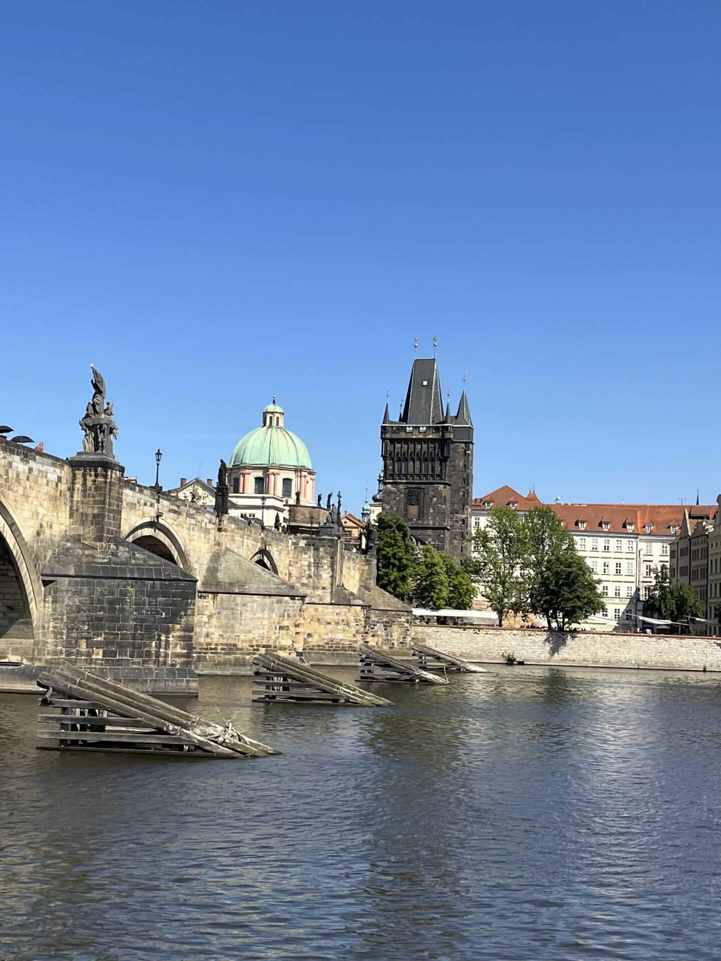 Several buildings line the Vltava river in Prague including a domed building with a green roof and a tall dark building with spires. The sky is clear blue overhead.