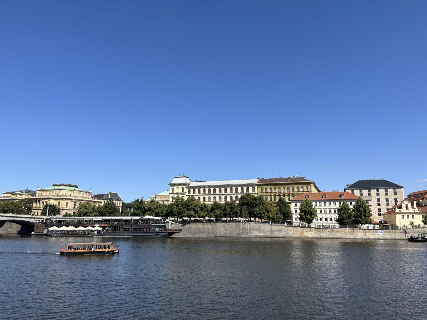 A wooden boat on the Vltava river, on the opposite bank there are several large historic buildings with a mix of domed and flat roofs. The buildings are surrounded by trees and greenery. The sky is clear and bright blue.