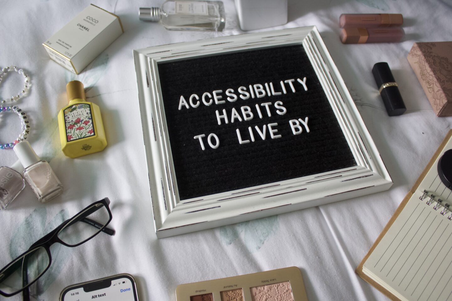 A flatlay arrangement with various items set out on a white surface. In the centre is a black felt letterboard with a white rustic frame and white letters that spell out Accessibility habits to live by. Several items surround the board including a pair of black glasses, a couple of perfumes, a phone displaying Instagram's ALT text page and a few makeup products.