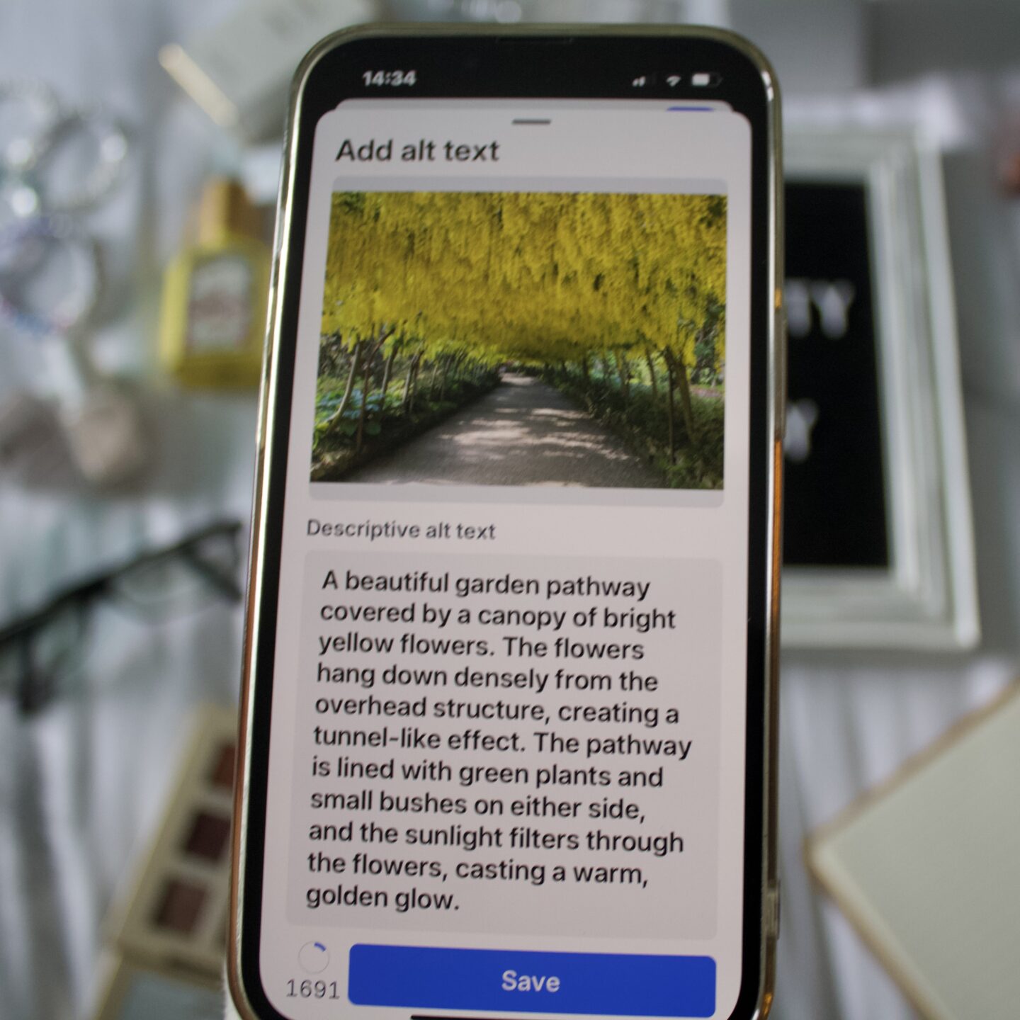 BlueSky's Add ALT text window displayed on a phone screen. A large box shows the descriptive text for the photo above. It says A beautiful garden pathway covered by a canopy of bright yellow flowers. The flowers hang down densely from the overhead structure, creating a tunnel-like effect. The pathway is lined with green plants and small bushes on either side and sunlight filters through the flowers, casting a warm golden glow. A blue button at the bottom of the screen says 'Save'.