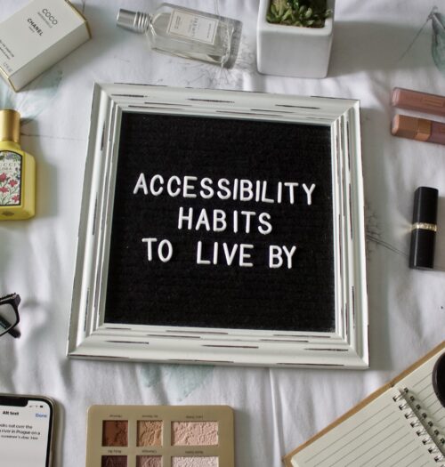 A black felt letterboard with a white rustic frame and white letters that spell out 'Accessibility habits to live by' is placed on a flat white surface with various items surrounding it, including a phone displaying Instagram's ALT text page, a notebook, a pair of black glasses, a few perfumes, a fake cactus and makeup products.