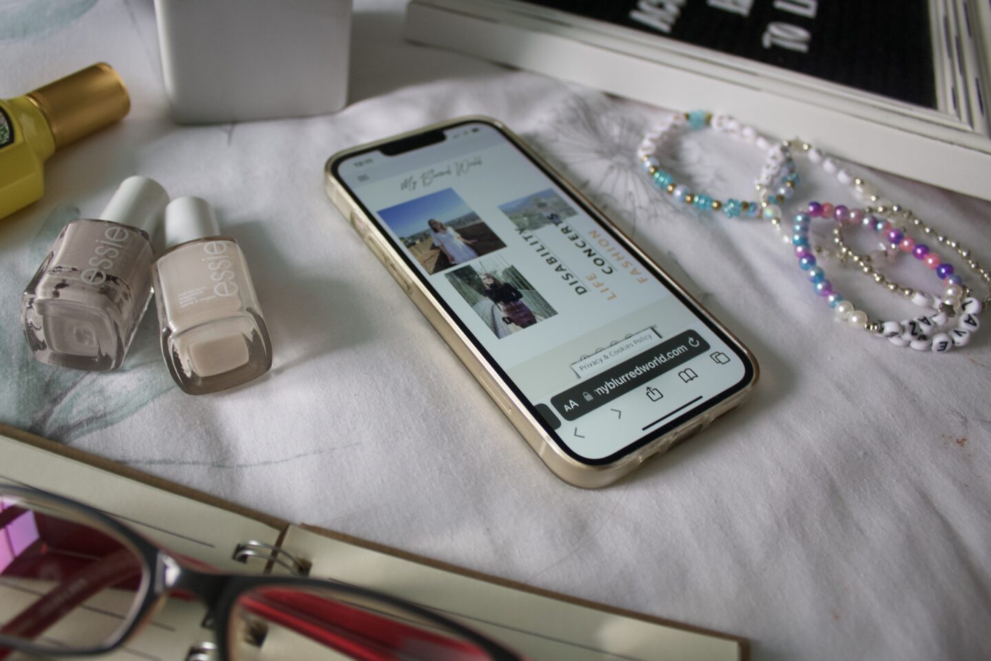 The My Blurred World blog homepage is displayed on a phone which is placed on a flat white surface and is surrounded by an open notebook with a pair of glasses resting on it, a couple of nail polishes and a perfume bottle.