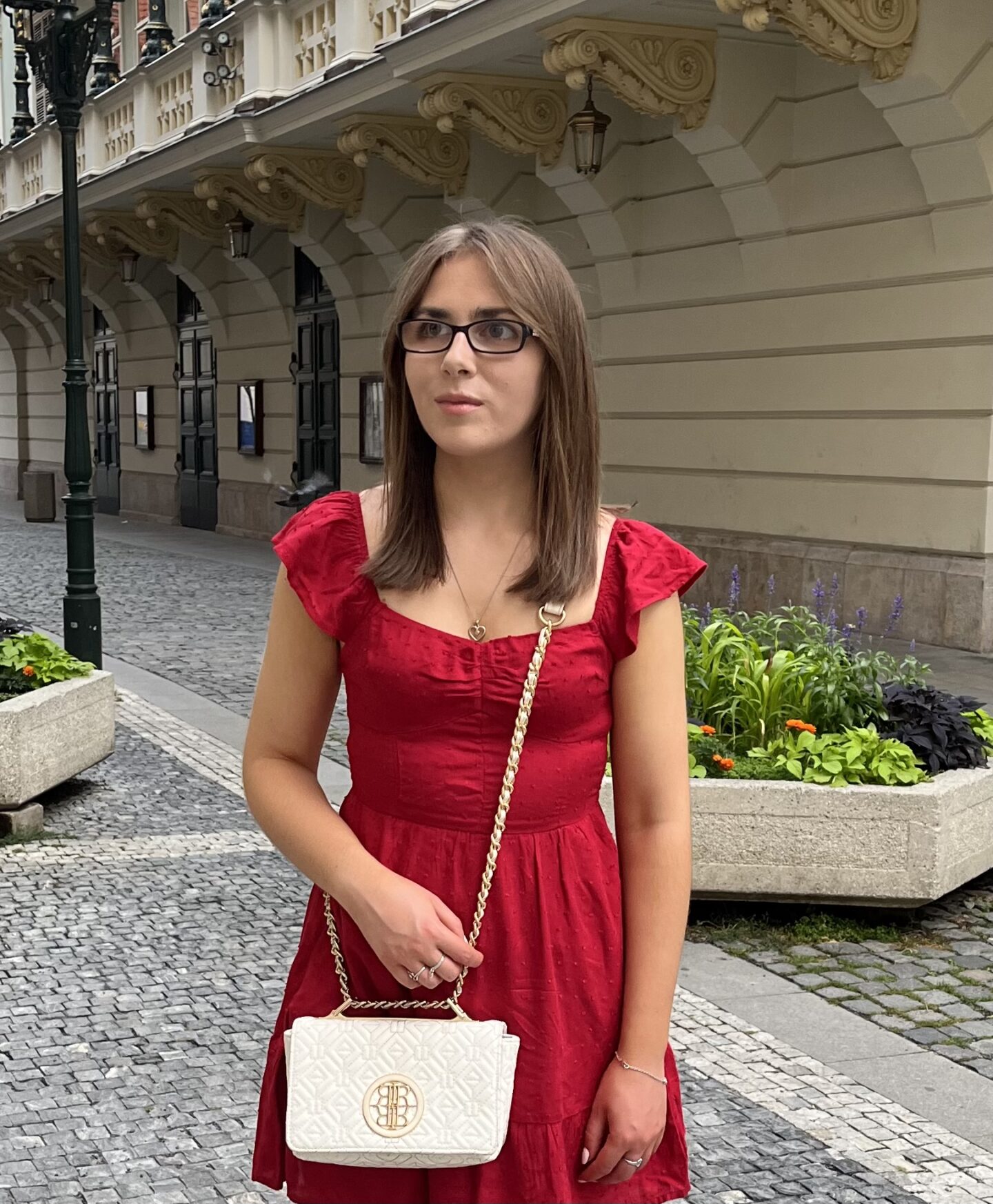 Elin standing on a cobblestone street in front of a large ornate building which has a row of green plant pots in front of it. She's looking away from the camera to her left.