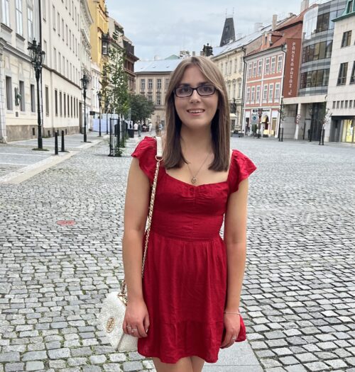 Elin standing on a cobble stone street in Prague. She wears a bright red mini dress with short flutter sleeves and a sweetheart neckline. She has long straight brown hair and is wearing glasses. She is carrying a small white handbag with gold hardware and a chain strap over her shoulder. The street is lined with historic buildings and a few lampposts. The sky is overcast and there are a few people in the background.