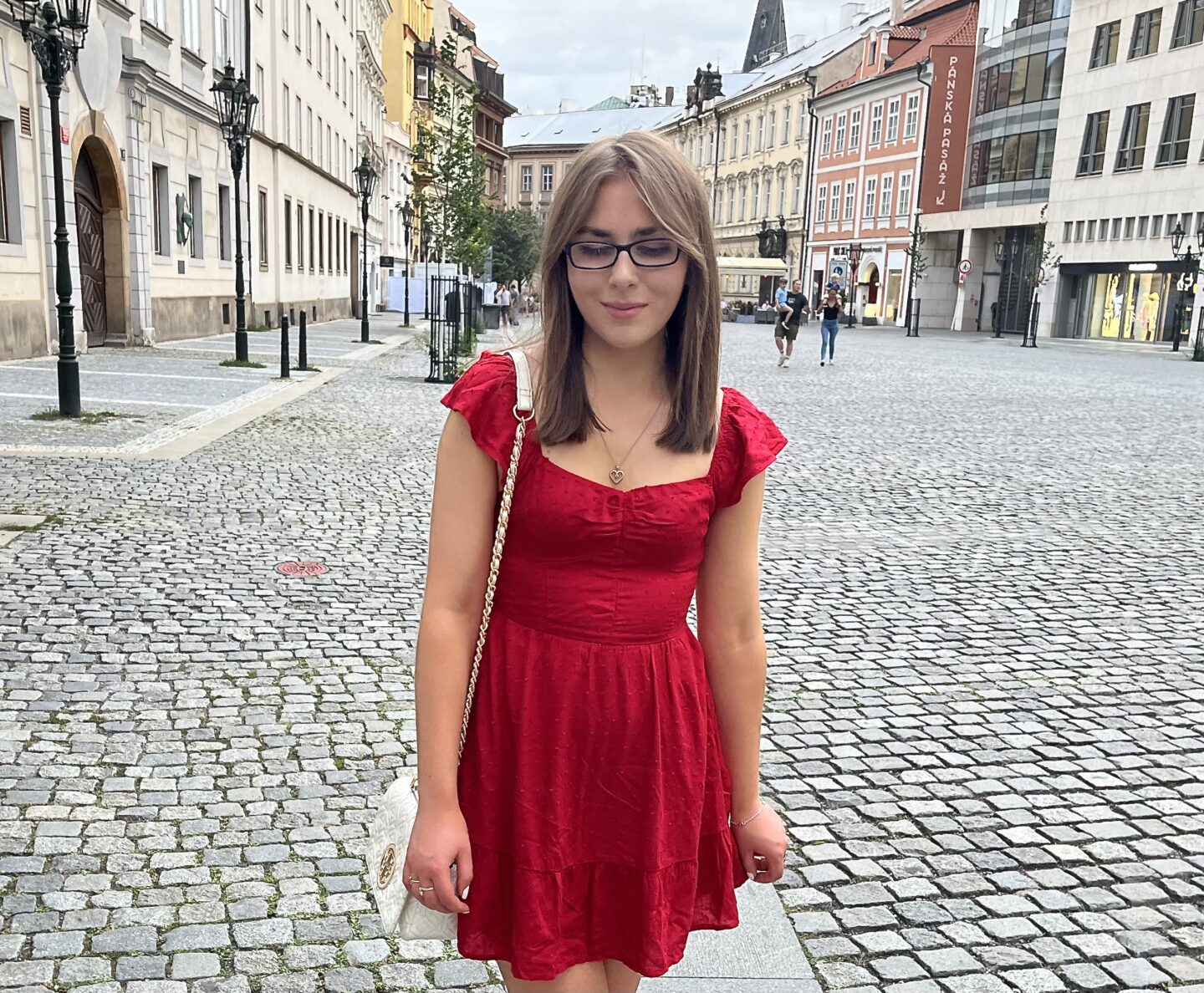 Elin standing on a cobblestone street with a mix of historic and slightly more modern buildings lining either side. She is looking down reflectively and is lightly holding the hem of her red mini dress which has short flutter sleeves and a sweetheart neckline. She's paired the number with a white bag which has gold hardware and a dainty heart shaped silver necklace.