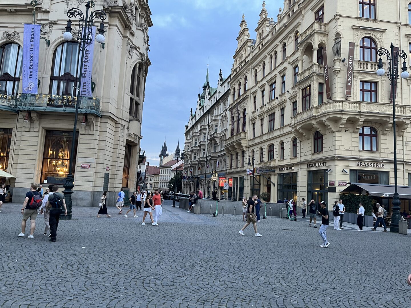 A busy street in Prague which is paved with cobblestones and is lined with large historic buildings on either side. A couple of signs on the buildings are for a brasserie and cafe.