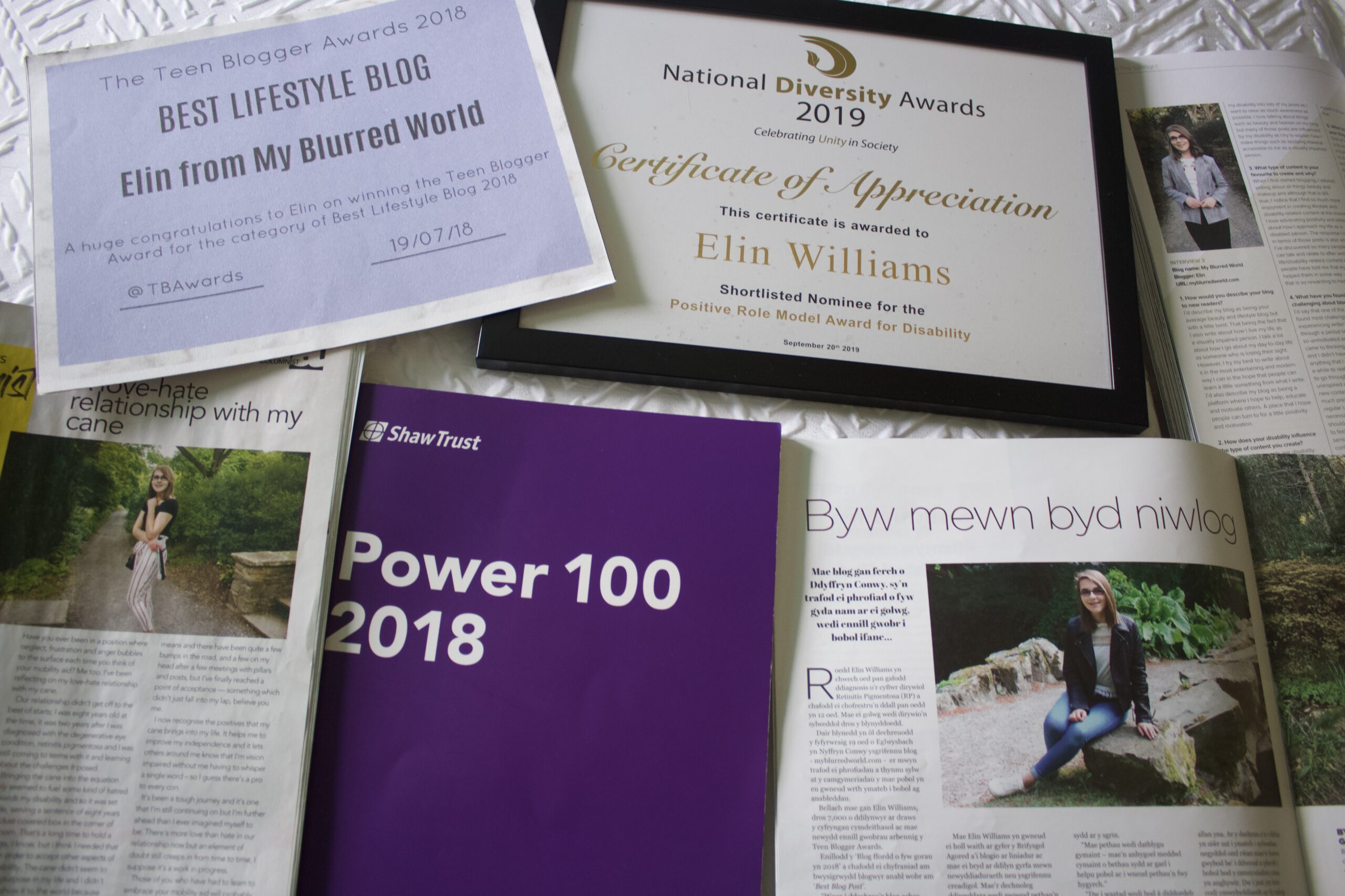 A certificate for the Teen Blogger Awards Best Lifestyle Blog of 2018, a framed certificate for being a shortlisted nominee for the Positive Role Model for Disability at the National Diversity Awards, the Shaw Trust Disability Power 100 List, an open copy of Blogosphere, Able, and Golwg magazines are spread out on a white textured surface. The magazines show interviews with Elin along with an article she wrote.