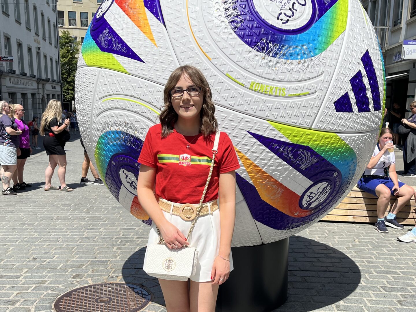 Elin standing in front of a giant decorative football sculpture. She's wearing a bright red t-shirt with a colourful horizontal stripe and a small emblem in the centre with the Wales flag and the Word Cymru in it. She's paired it with white high waisted tailored shorts, a raffia belt and a white crossbody bag. The ball behind her is white with textured patterns and is decorated with bold colourful geometric designs in purple, blue, green, yellow and orange, as well as logos including the UEFA Women's Euros logo.