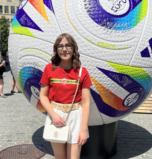 Elin standing in front of a giant decorative football sculpture. She's wearing a bright red t-shirt with a colourful horizontal stripe and a small emblem in the centre with the Wales flag and the Word Cymru in it. She's paired it with white high waisted tailored shorts, a raffia belt and a white crossbody bag. The ball behind her is white with textured patterns and is decorated with bold colourful geometric designs in purple, blue, green, yellow and orange, as well as logos including the UEFA Women's Euros logo.