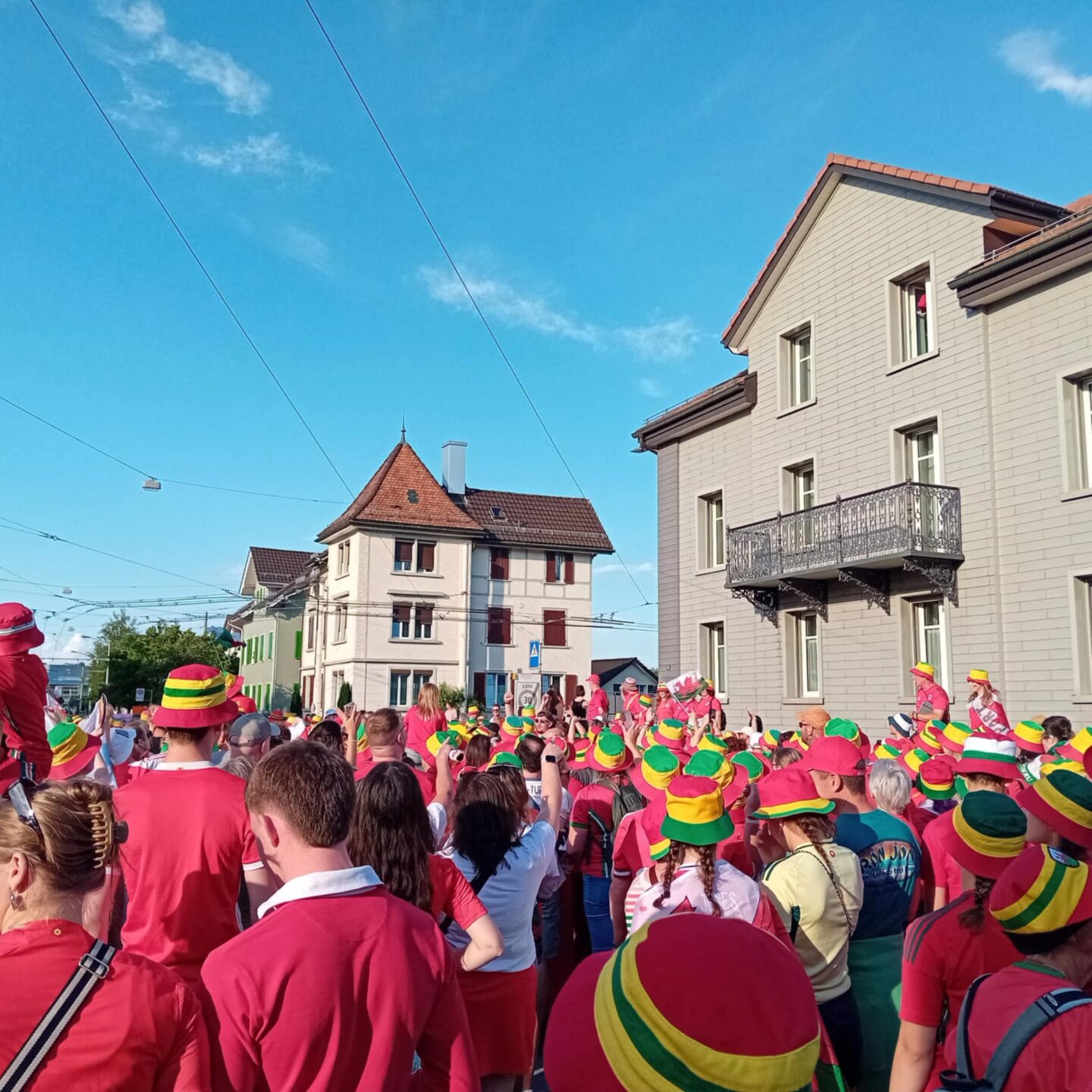A crowd of people moving through St. Gallen as part of the fan walk. Most are wearing red tops and bucket hats striped with red, yellow and green. The street is lined with European style buildings, characterised by light facades, multiple windows and sloped roofs with red tiles.