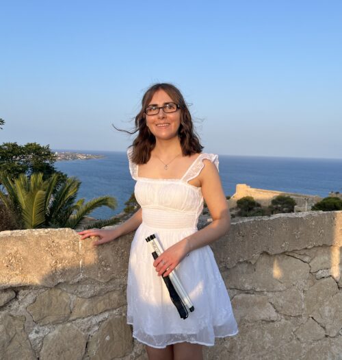 Elin standing by a stone wall with her right hand resting on it. She wears a white mini dress with embroidered details, a square neckline and sheer flutter sleeves. She holds her white cane in her left hand. A mix of scenery is in the background from the tall buildings of a city to greenery and the sea.