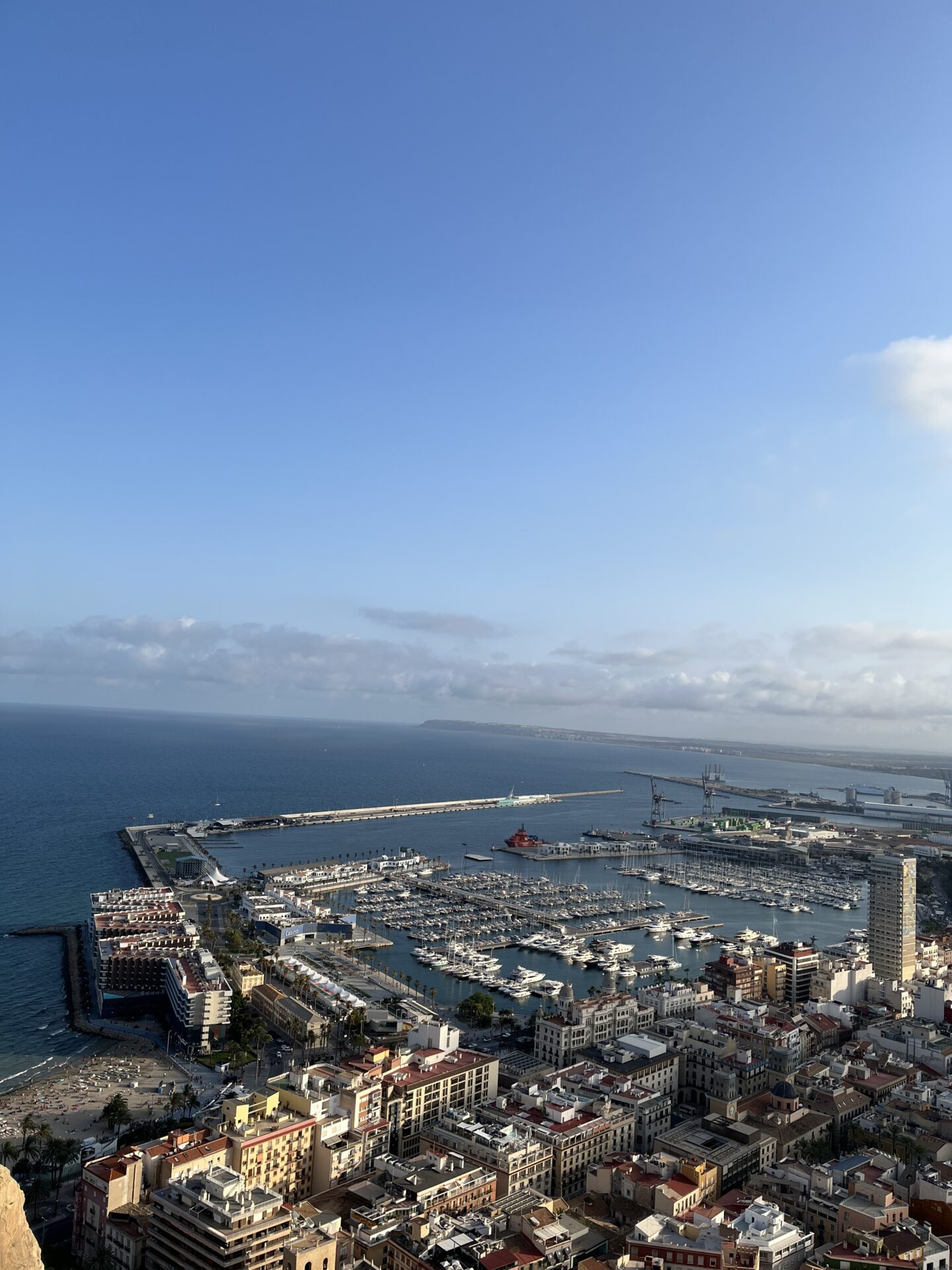 A view of Alicante from the elevated castle walls. There is a marina filled with boats, densely packed buildings, and a pier jutting out into the blue sea. There is a sandy beach on the left side.