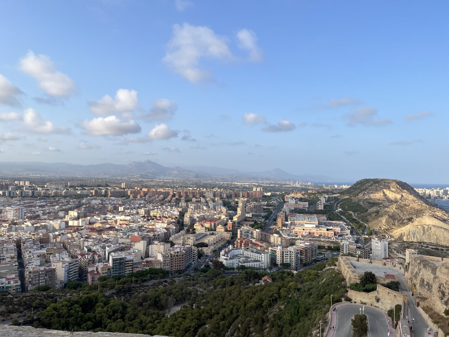 A cityscape viewed from the elevated castle walls. There are densely packed buildings, streets and greenery. In the foreground, there are green trees and a winding road leading up a hill. To the right, a rocky hill rises above the city. The city stretches out in the distance with mountains slightly visible on the far horizon.