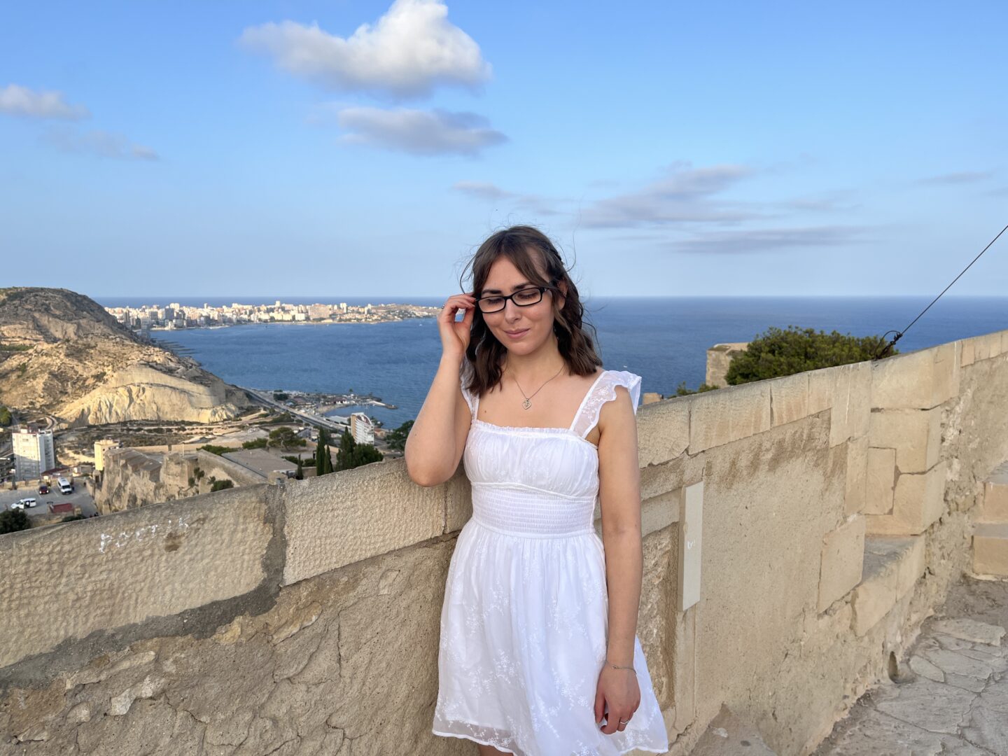Elin standing by a stone wall which overlooks a view of Alicante City with its old buildings, rocky hills and coastline. Her right hand is raised to hold a strand of her curled hair away from her face and she’s looking down. She’s wearing a white mini dress with subtle embroidered details, black framed glasses and silver jewellery.