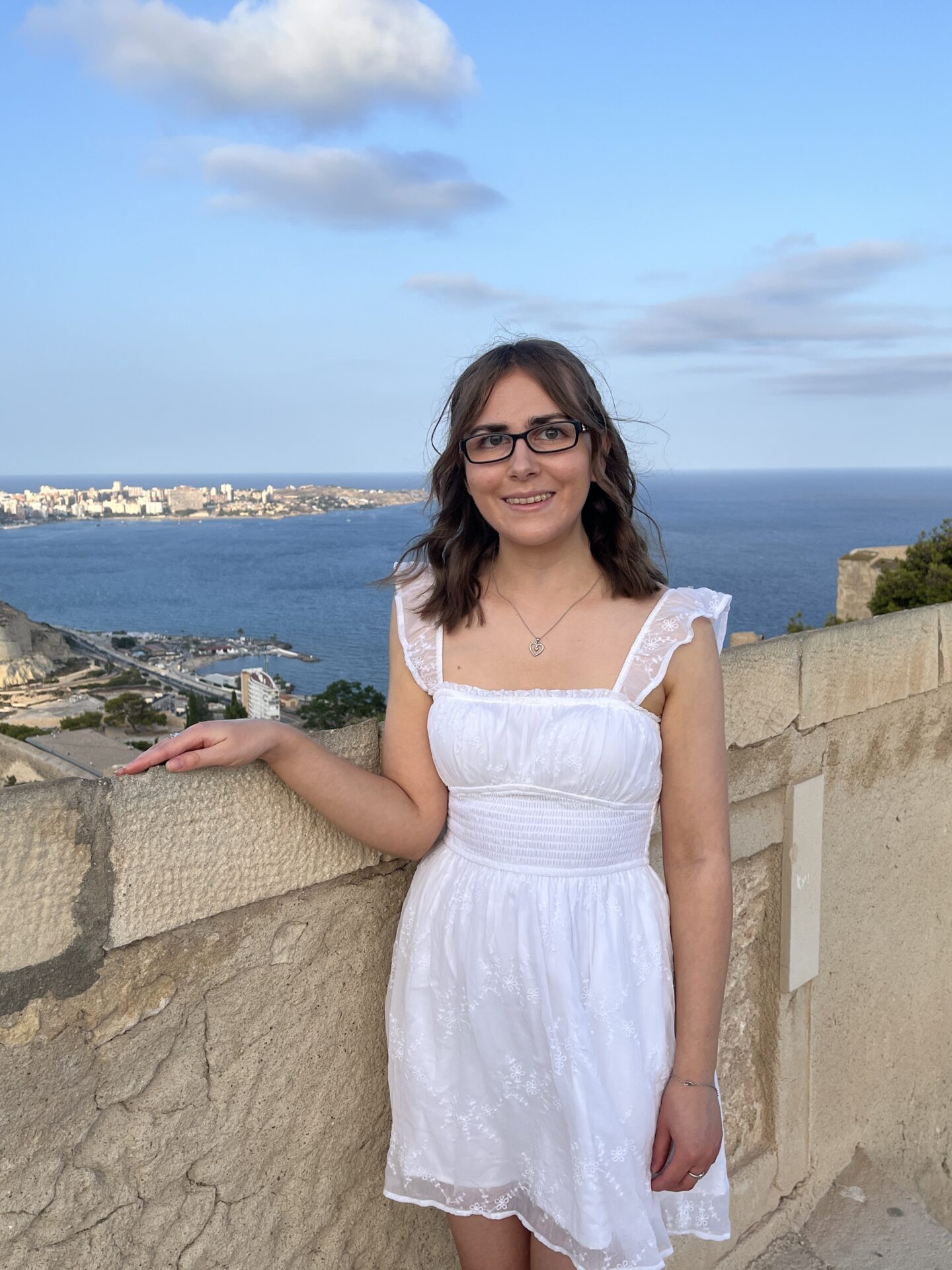 Elin standing in a white mini dress by a stone wall with her right hand resting atop it. The background shows the sea with a cityscape and coastline visible to the right. She’s smiling at the camera.