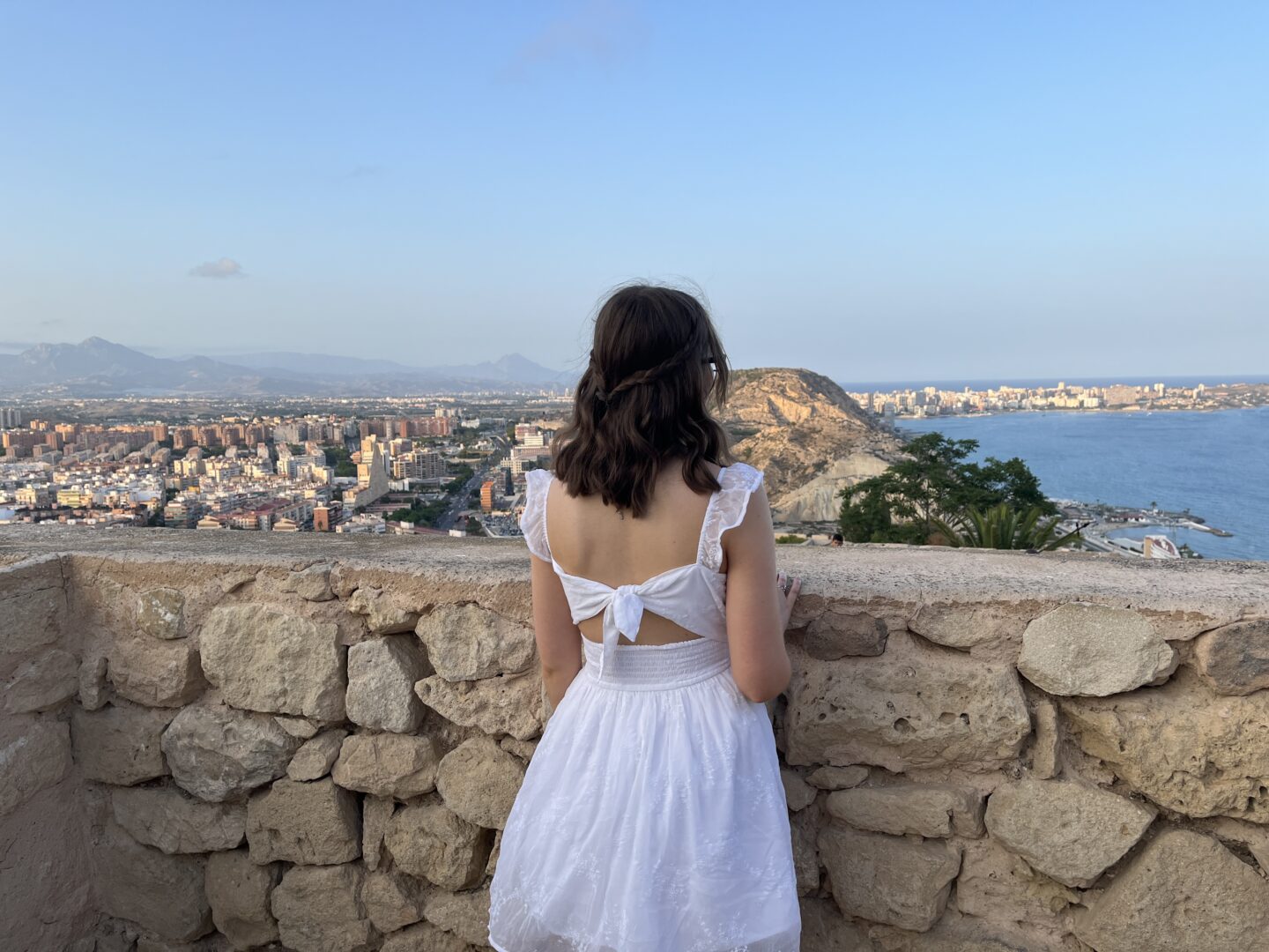 Elin standing with her back facing the camera, overlooking a coastal view of Alicante from the elevated castle walls. She’s wearing a white mini dress with a white bow tied at the back and embroidered details. Her hair is styled into a curly half-up, half-down do with two braids tied together at the back. The view consists of the city's buildings, a large hill and the blue sea stretching out to the right. The sky is clear and blue with distant mountains visible on the left.