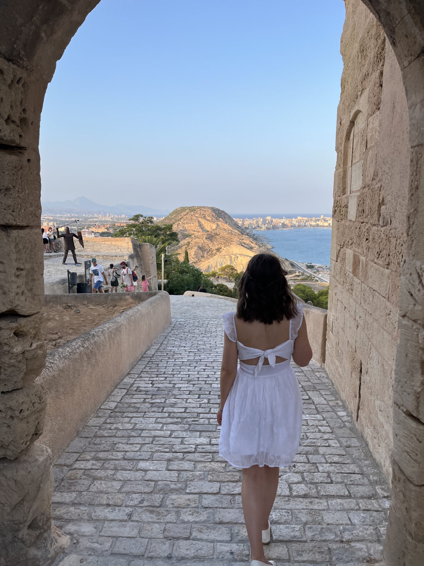 Elin walking down a cobblestone path through a stone archway. The path leads toward a scenic view of Alicante City bordered by a calm blue sea. Her white embroidered mini dress has an open back with two pieces of fabric tied in a bow. It has sheer flutter straps with a fitted bodice which connects to a flared skirt. There are a few other people dotted around the viewpoint.