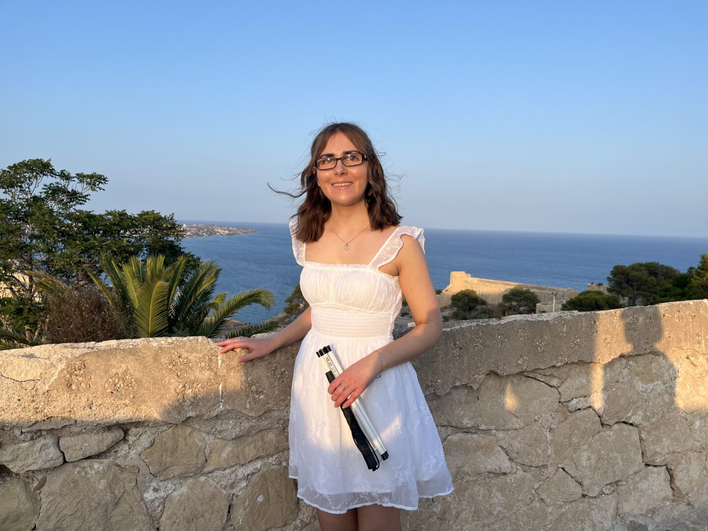 Elin standing by a stone wall with her right hand resting atop it. Her folded up cane is in her left hand. She’s wearing a white mini dress which has subtle matching embroidery all over, a square neckline, and sheer flutter straps. Her hair is styled into a curled half-up, half-down do but the wind has caught some strands. It’s golden hour and there is a scenic coastal view behind her, showing palm trees, greenery and the bright blue sea under a clear sky. She’s smiling at the camera.
