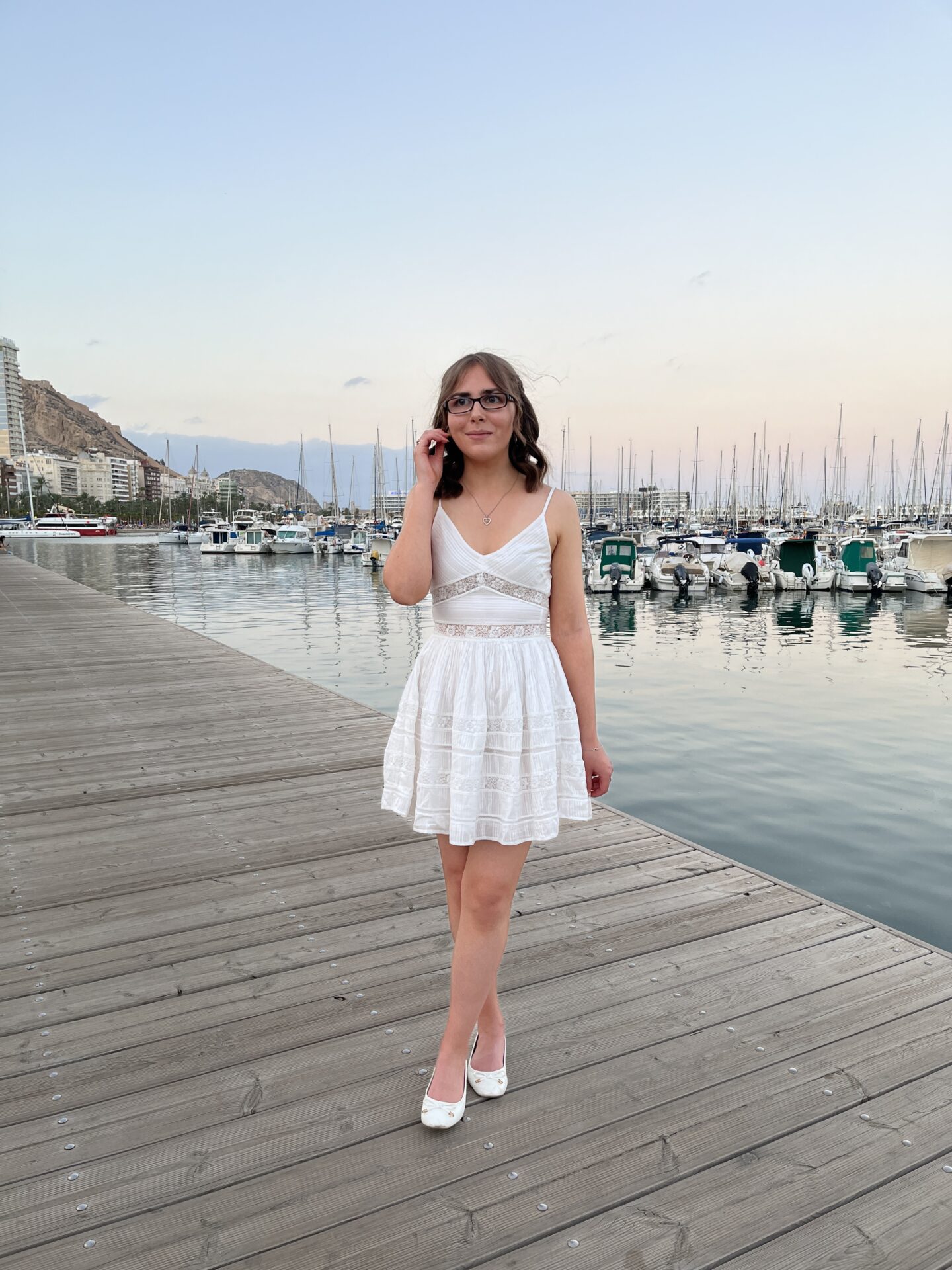 Elin standing on a wooden dock by a marina which is filled with boats and yachts. The water reflects their masts and some lights from the buildings on the shore. Elin's looking away from the camera to her right.