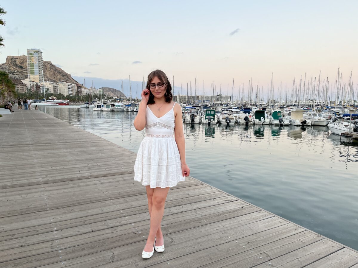Elin standing on a wooden dock by a marina which is filled with boats and yachts. She's wearing a white mini dress with lace inserts at the waist and embroidered details all over. It has a v-neckline and spaghetti straps. Her brown hair is styled into a half-up, half-down do and her right hand is raised to push a strand away from her face. She's looking down reflectively. In the background is a rocky hill with Alicante's Santa Barbara castle slightly visible at the top.