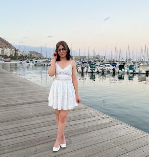 Elin standing on a wooden dock by a marina which is filled with boats and yachts. She's wearing a white mini dress with lace inserts at the waist and embroidered details all over. It has a v-neckline and spaghetti straps. Her brown hair is styled into a half-up, half-down do and her right hand is raised to push a strand away from her face. She's looking down reflectively. In the background is a rocky hill with Alicante's Santa Barbara castle slightly visible at the top.