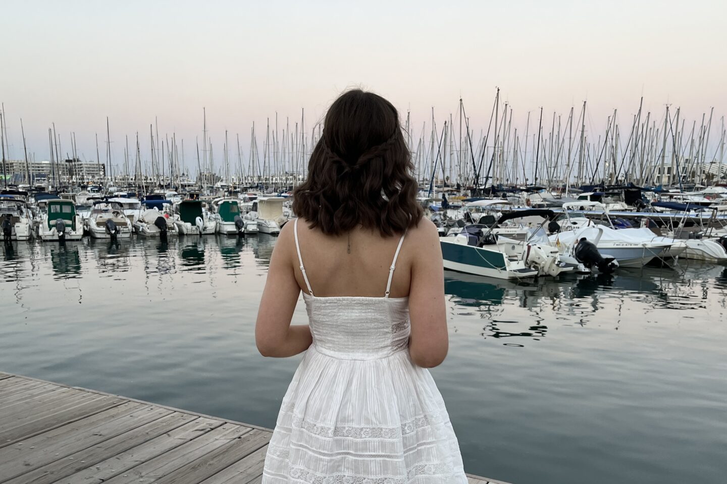 Elin standing on a wooden dock, facing away from the camera. She wears a white mini dress with spaghetti straps and her brown hair is styled into loose curls with two braids tied together at the back. She's looking out at several boats and yachts docked at the marina.