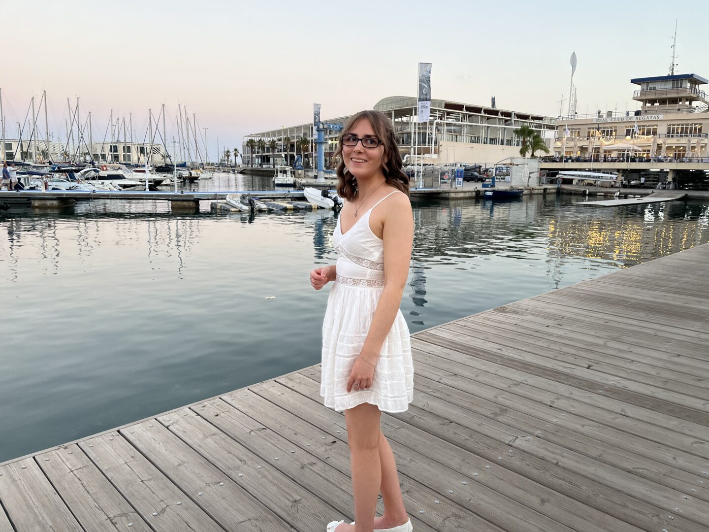 Elin standing on a wooden dock by a marina. She's smiling broadly at the camera and wears a white mini dress with a lace insert at the waist and embroidered details all over. Several boats are docked on the water which reflects the sky's slightly pastel hues and the lights of the buildings on the shore.