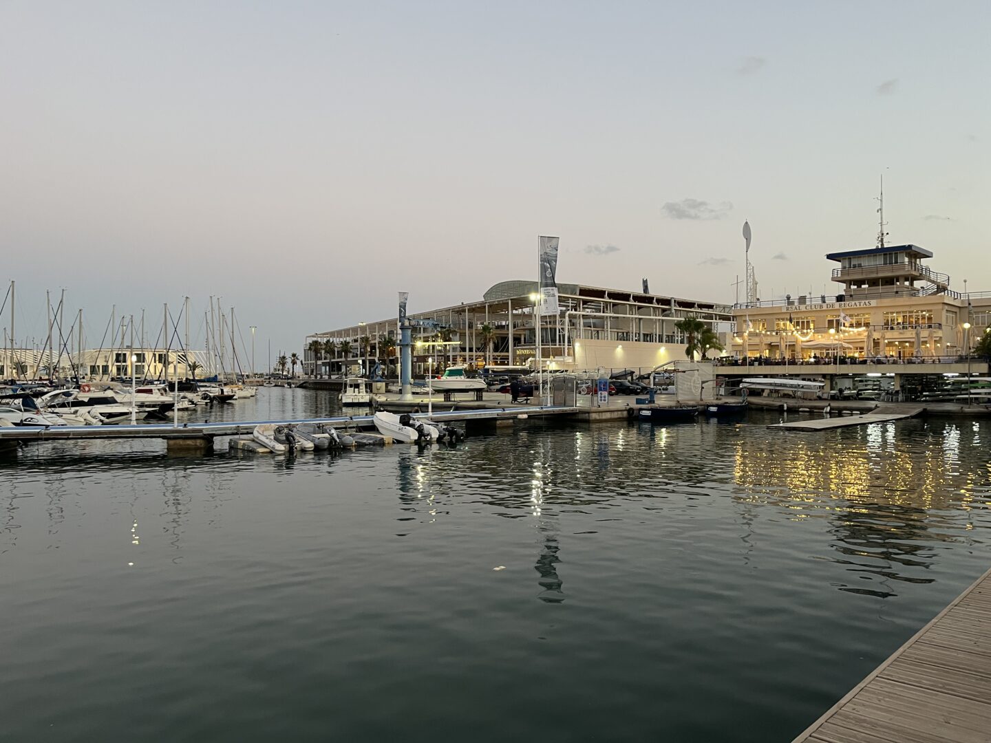A marina at sunset with calm water reflecting the lights from buildings on the shore. Several boats and yachts are docked with their masts standing tall.