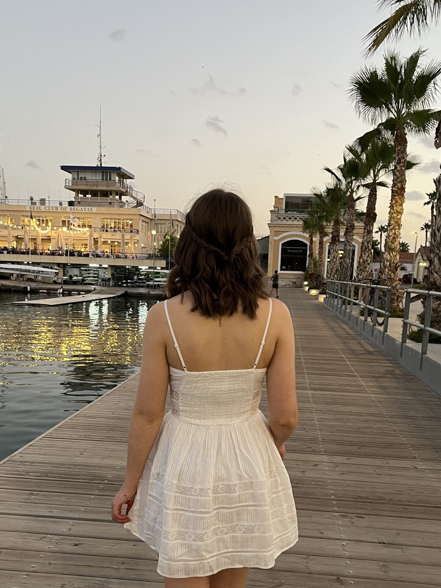 Elin walking away from the camera on a wooden boardwalk. Her medium length brown hair is styled into loose waves with two braids tied together at the back. There are several boats docked on the water on the left and there are palm trees on the right.