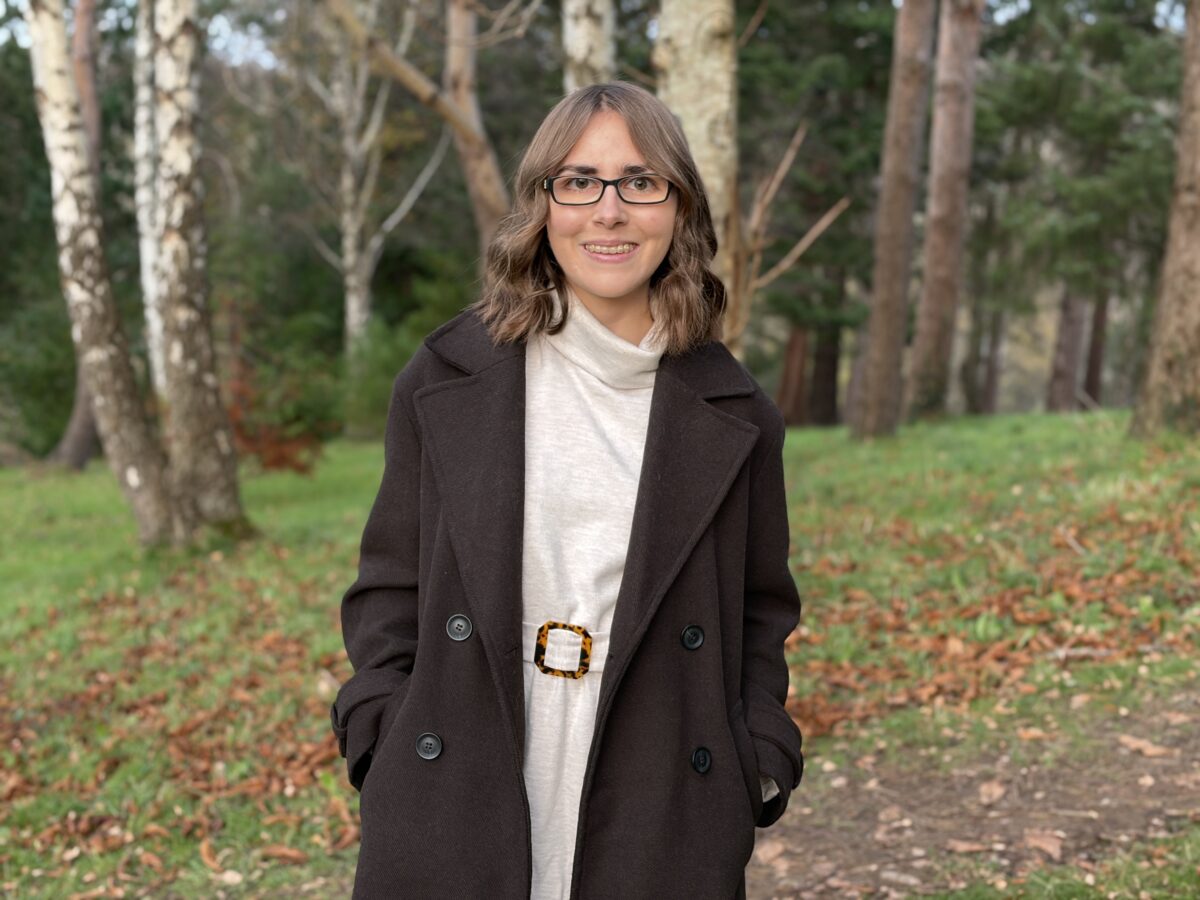 Elin standing in the woods with fallen leaves on the ground all around her. She's wearing a chocolate brown double breasted coat over a cream belted jumper dress which has a roll neck. Her hands are in her coat pockets and she's smiling broadly at the camera. Her brown hair is styled into loose waves which hang just above her shoulders.