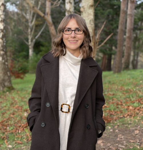 Elin standing in the woods with fallen leaves on the ground all around her. She's wearing a chocolate brown double breasted coat over a cream belted jumper dress which has a roll neck. Her hands are in her coat pockets and she's smiling broadly at the camera. Her brown hair is styled into loose waves which hang just above her shoulders.