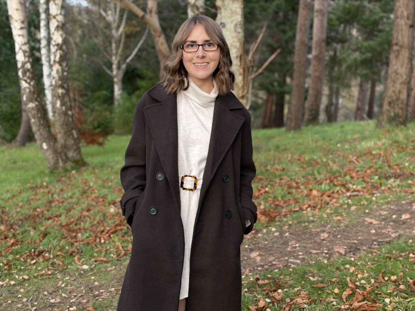 Elin standing in the woods with fallen leaves on the ground all around her. She's wearing a chocolate brown double breasted coat over a cream belted jumper dress which has a roll neck. Her hands are in her coat pockets and she's smiling broadly at the camera. Her brown hair is styled into loose waves which hang just above her shoulders.