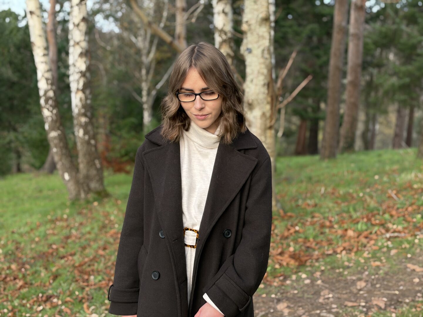 Elin standing in the woods with fallen leaves scattered on the ground all around her. She's looking down reflectively.