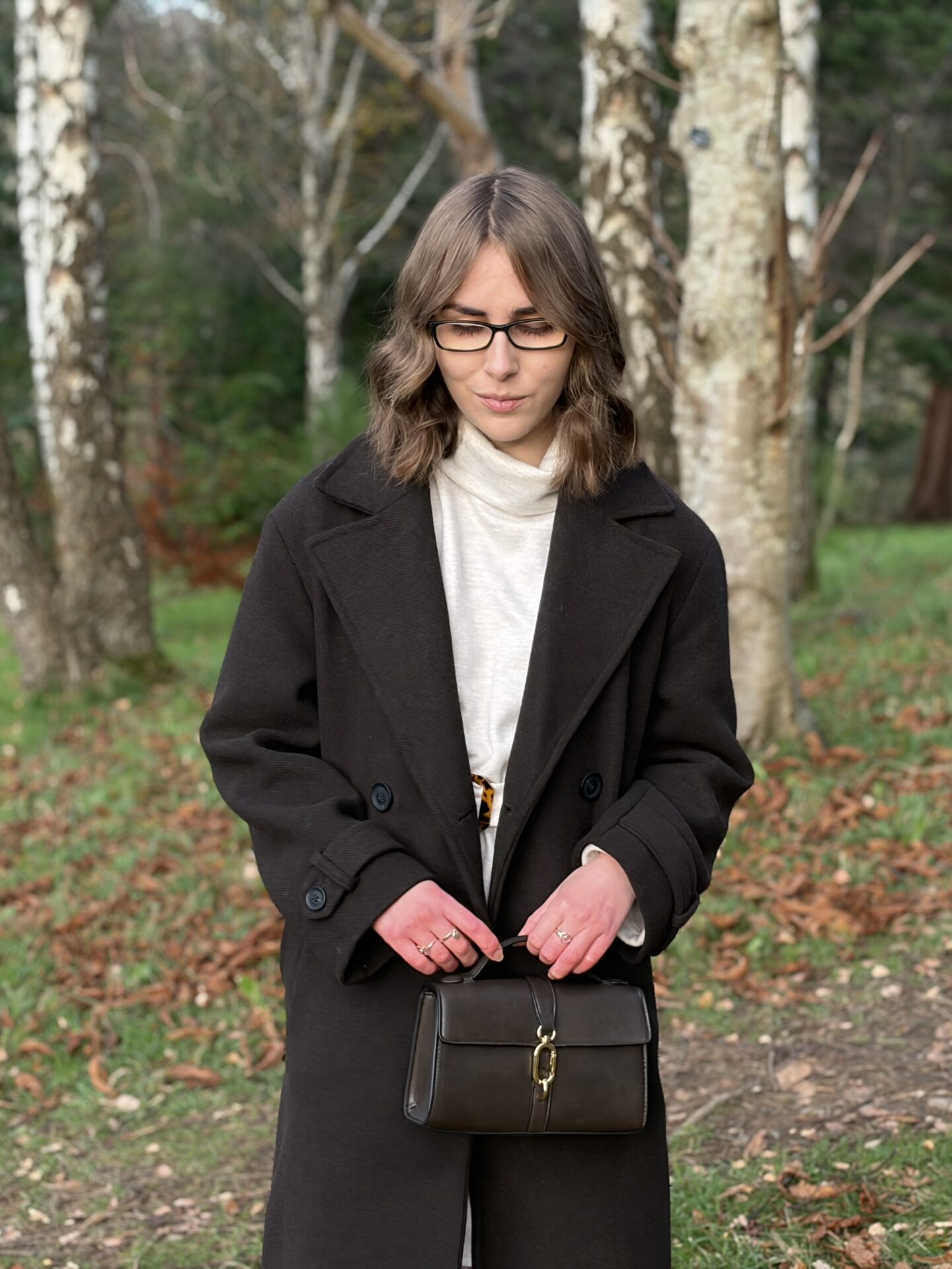 Elin looking down reflectively. She's holding the handle of a small dark brown handbag with a gold clasp with both her hands in front of her.
