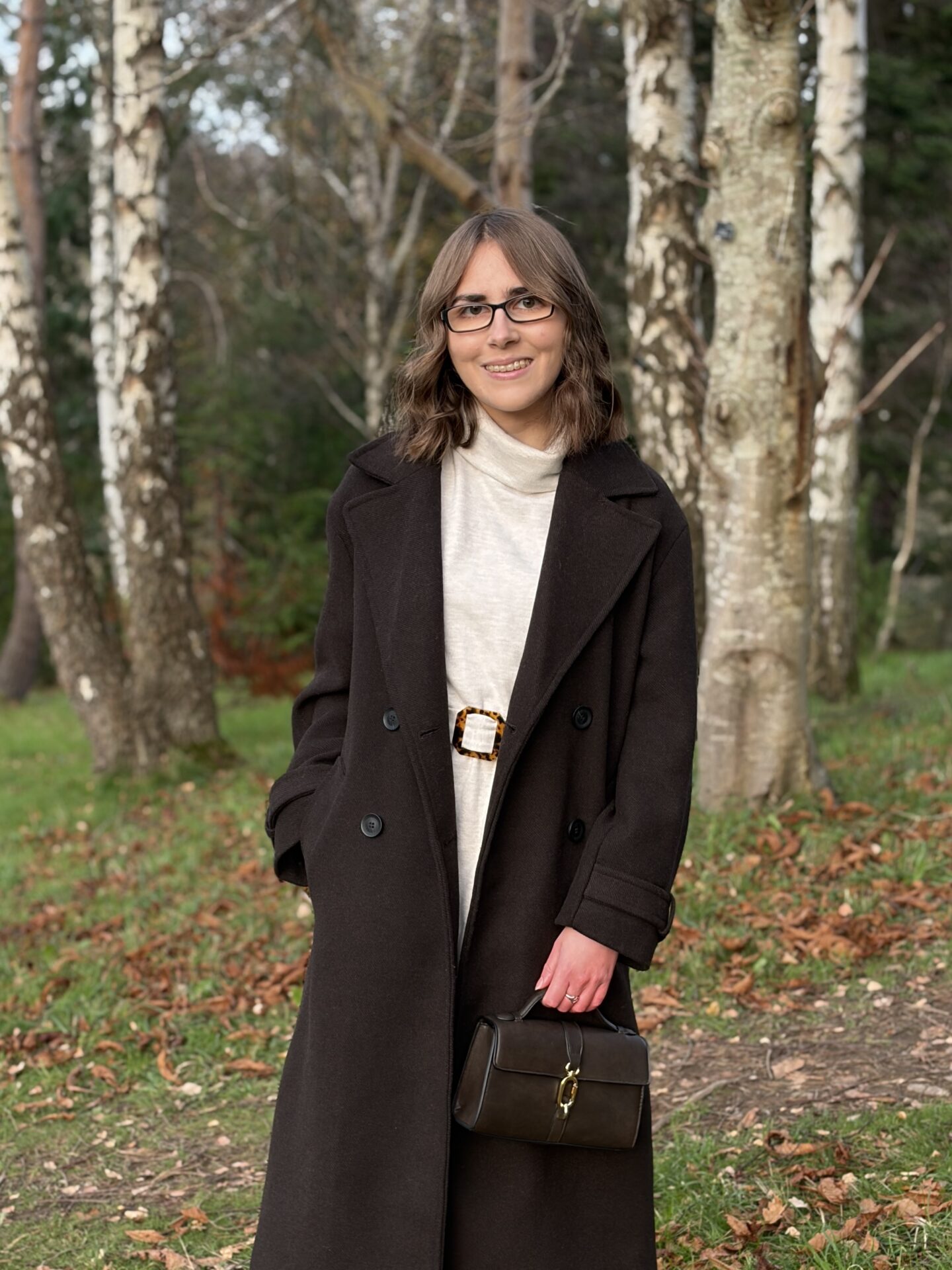 Elin standing in the woods. She wears a brown longline coat over a cream jumper dress and is holding a small brown handbag which has a gold buckle fastening in her left hand. She's looking at the camera and has a soft smile.