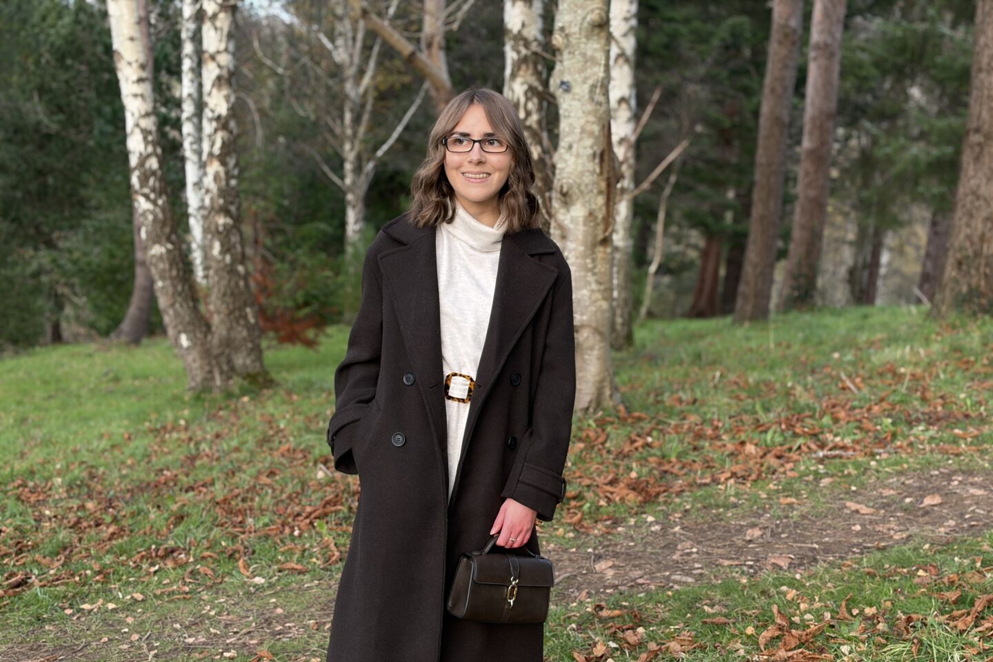 Elin looking away from the camera and smiling. She's standing in woodland and wears a long brown coat over a cream jumper dress. She's holding a small dark brown handbag in her left hand. Her brown hair is styled into loose waves.