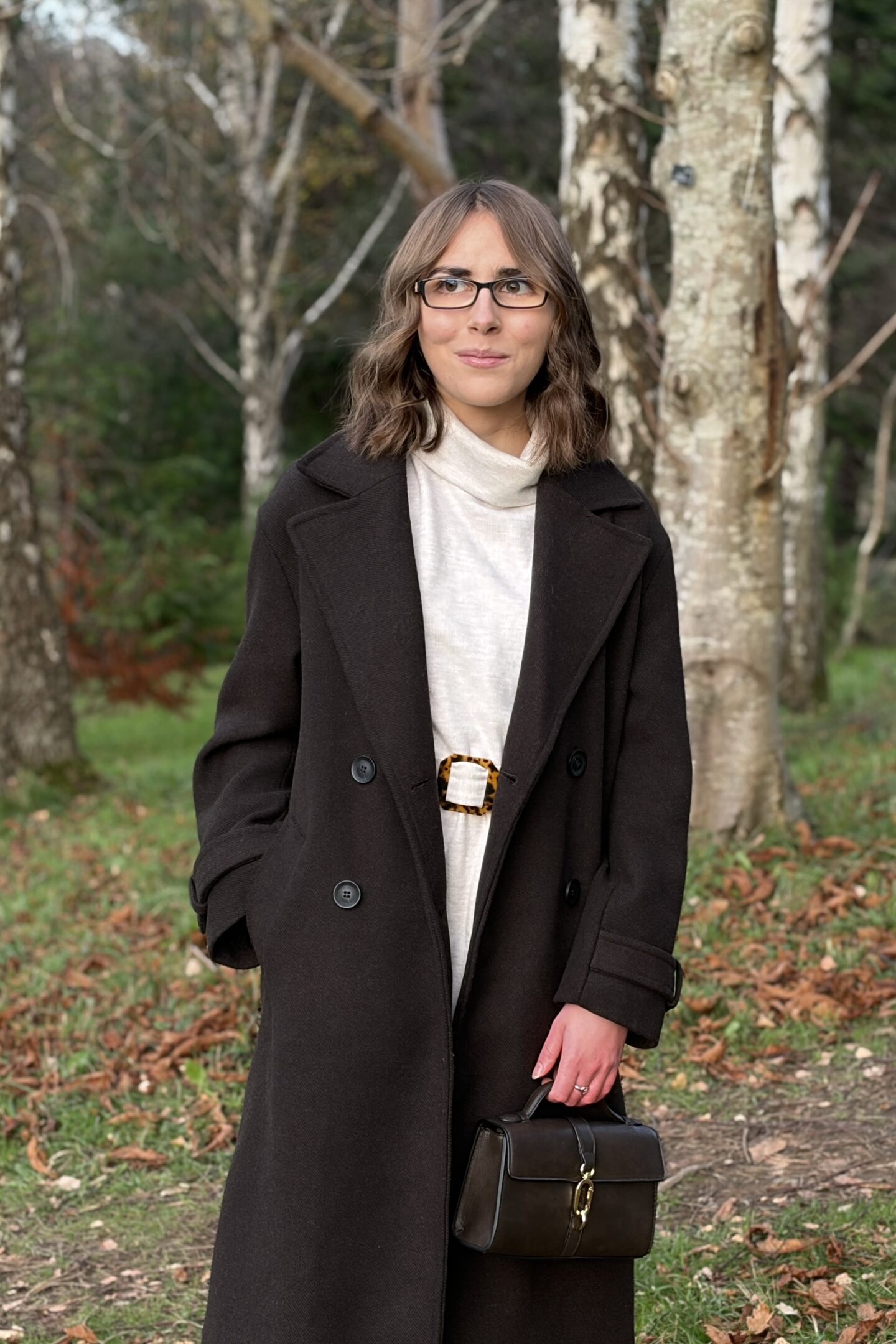 Elin standing in woodland, looking away from the camera. She's styled her brown hair into loose waves and is wearing a brown coat over a cream jumper dress which has a roll neck. She's carrying a small brown handbag in her left hand.