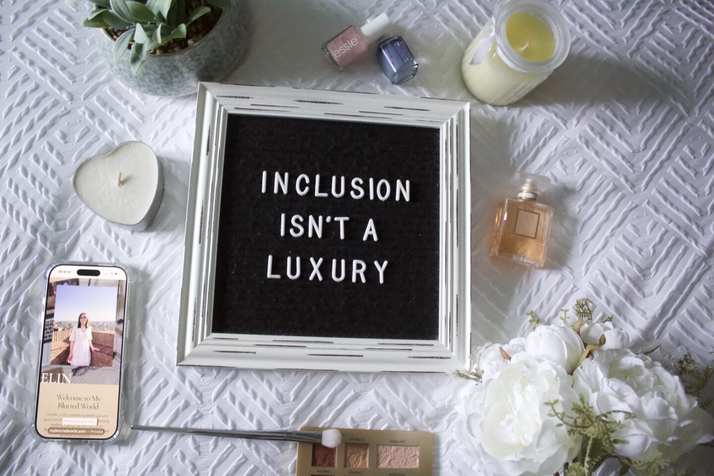 A black felt letter board with a retro white wooden frame and white letters reads: Inclusion isn't a luxury. The board is set on a textured white blanket and is surrounded by a fake plant, fake white peonies, two candles, a couple of nail polishes, a small perfume bottle and a phone which displays the My Blurred World blog homepage.