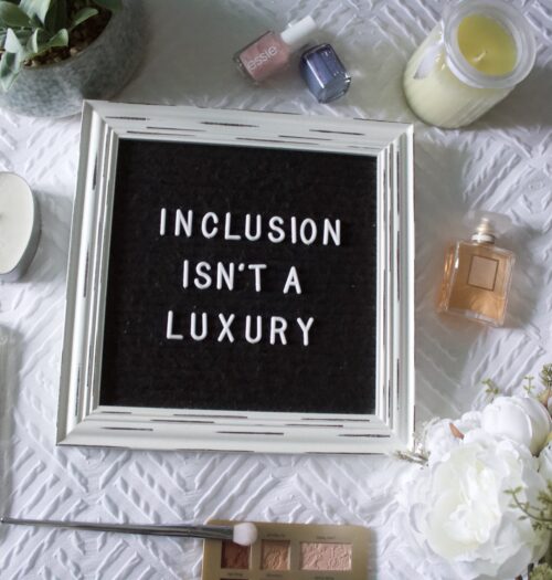 A black felt letter board with a retro white wooden frame and white letters reads: Inclusion isn't a luxury. The board is set on a textured white blanket and is surrounded by a fake plant, fake white peonies, two candles, a couple of nail polishes, a small perfume bottle and a phone which displays the My Blurred World blog homepage.