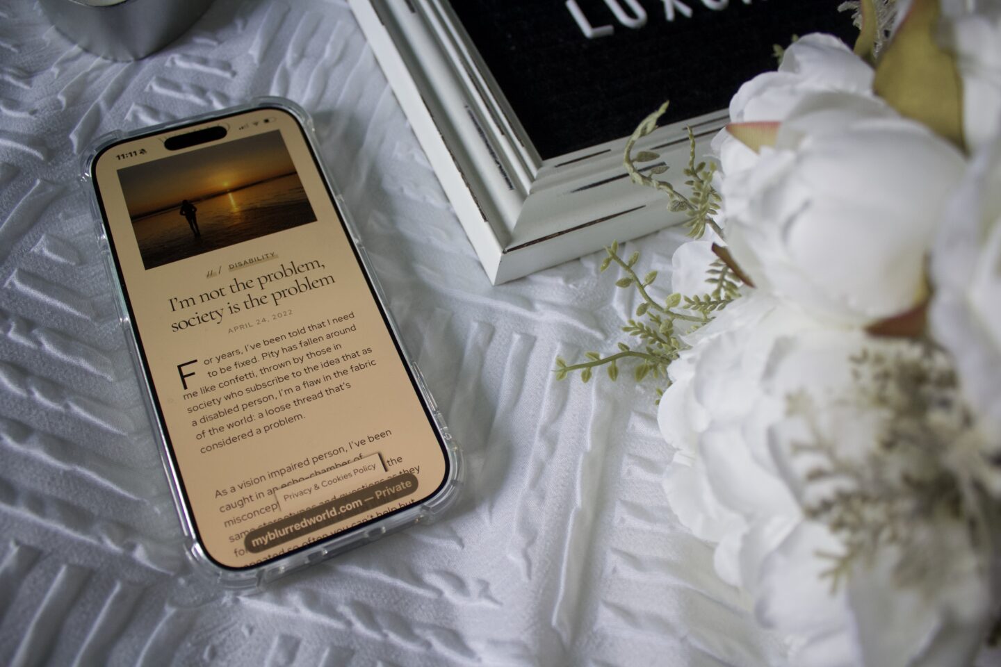 A phone displays a blog post titled I'm not the problem society is the problem. The phone is set on a white textured blanket beside a black and white letter board which is slightly visible in the shot along with a bouquet of artificial white peonies.