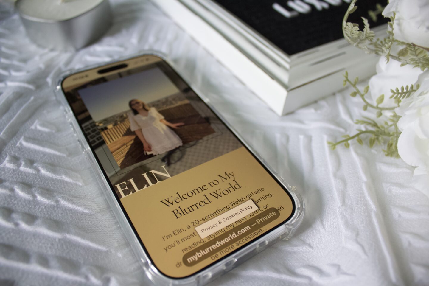 A phone displays the My Blurred World blog homepage. The phone is set on a white textured blanket beside a black and white letter board which is slightly visible in the shot along with a bouquet of artificial white peonies and a small heart shaped candle.