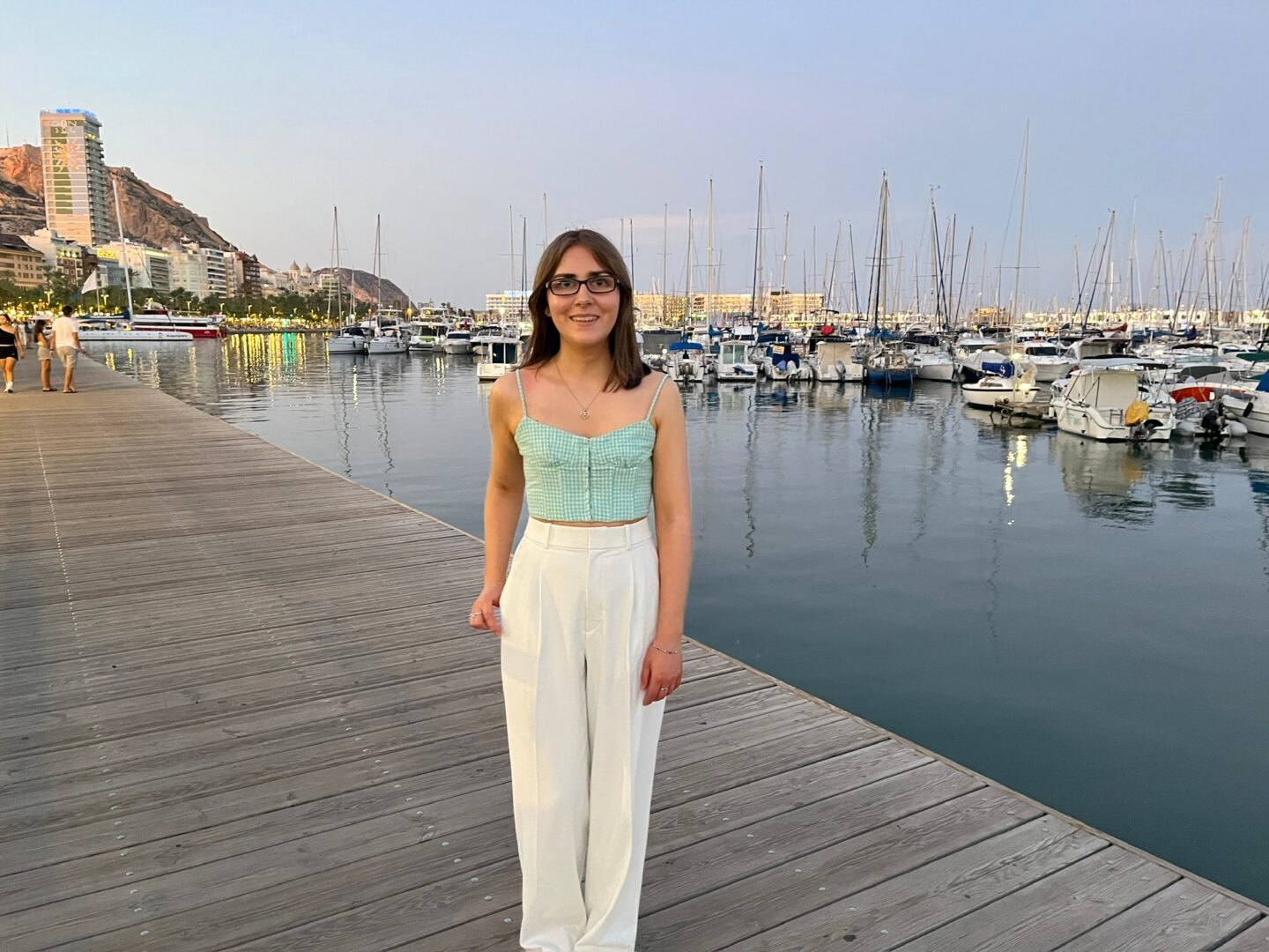 Elin standing on a wooden boardwalk by a marina at dusk. She wears a gingham green and white crop top with thin straps, white high waisted wide leg trousers with a subtle pleated detail and white quilted ballet flats. Her right thumb is hooked into her pocket and she's smiling broadly at the camera.