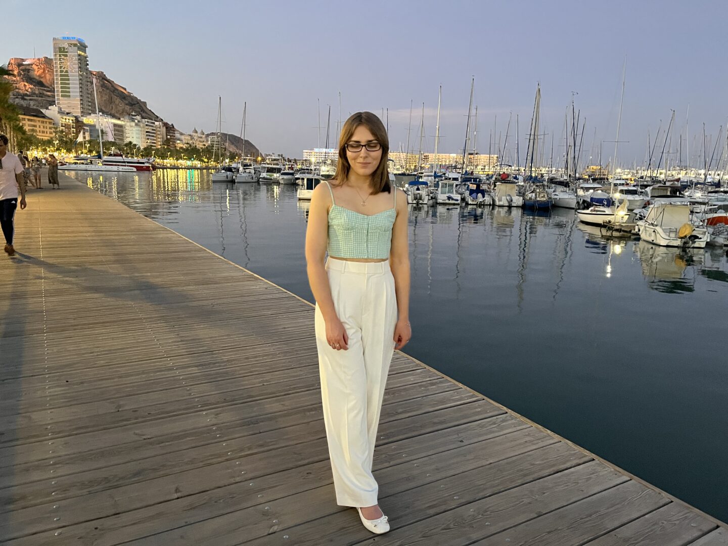 Elin standing on a wooden boardwalk by a marina at dusk. She has straight shoulder length brown hair and wears a light green gingham crop top with white pleated trousers. Her arms are relaxed by her sides and she's looking down with a slight smile. In the distance is a rocky hill with Alicante's Santa Barbara castle atop it.