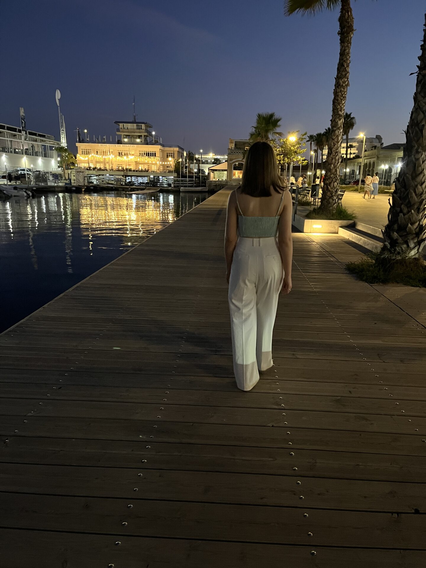 Elin walking away from the camera along a wooden boardwalk which stretches into the distance. The path is bordered by water on the left with several boats docked on it and lights from the shore shimmering on the surface. On the right side are tall palm trees and lit up buildings.