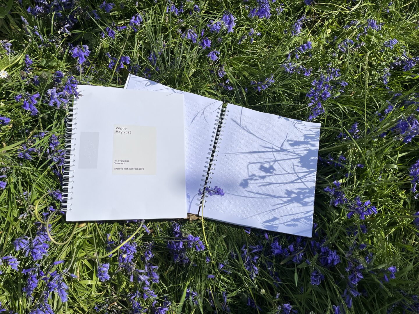 Two spiral bound braille editions of Vogue magazine lying on green grass scattered with bluebells. The magazine on the left is closed with a label on the front reading Vogue May 2023. This volume is placed on one side of the second volume which is open to show the raised braille dots inside.